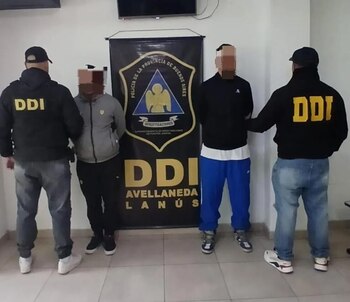 Cuatro hombres, dos con chaquetas negras DDI, flanquean a dos hombres con rostros pixelados, parados frente a un estandarte de la Policía de Buenos Aires