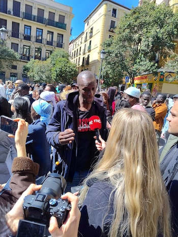 Serigne Mbaye, un hombre negro, habla a un micrófono rojo de RNE, sostenido por una mujer rubia. Otra persona graba con una cámara profesional. Fondo de multitud y edificios