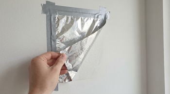 Primer plano de una mano despegando papel aluminio adherido con cinta a una pared blanca, mostrando gotas de agua condensada en la superficie interior del aluminio.