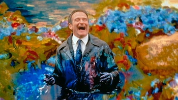 Un hombre de mediana edad con saco y corbata, cubierto de pintura azul y roja, sonríe y ríe con la boca abierta, rodeado de un fondo de colores brillantes