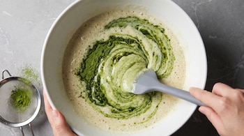 Manos sostienen un cuenco blanco con una mezcla clara y té matcha en polvo revuelto con una espátula gris, junto a un colador metálico.