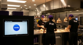 Pantalla digital Omilia en primer plano. Tres empleados de Taco Bell con gorras y uniformes preparan alimentos en una cocina con equipos de acero.