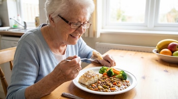 Mujer mayor con cabello blanco y gafas sonríe mientras come un plato de pescado, brócoli, zanahorias y lentejas en una mesa de madera. Se ve una ventana al fondo.