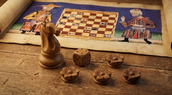Caballo de ajedrez, dado y cuatro piezas florales de madera en mesa rústica. Fondo: pergamino con ilustración de tablero de ajedrez y figuras.