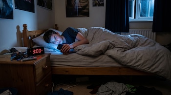 Adolescente masculino en la cama, iluminado por la pantalla de su celular, mientras un reloj digital marca las 3:00 AM en su mesita de noche.