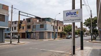 Un cartel con el texto Pluz Energía y Corte Programado en un poste de una calle con semáforos apagados y viviendas de varios pisos.