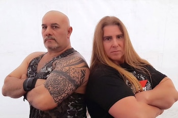 Dos hombres posan espalda con espalda, con los brazos cruzados. Uno calvo con barba, tatuajes y musculosa oscura. El otro con cabello rubio largo y camiseta negra. Fondo blanco