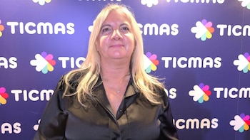 Primer plano de una mujer de cabello rubio claro con blusa negra y collar, sonriendo ligeramente frente a un fondo azul con el logo de Ticmas repetido