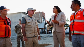 Cinco personas al aire libre, tres hombres con chalecos de emergencia y gorras, un militar y una mujer con traje sastre, están conversando. Edificios al fondo