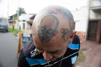 Close da cabeça de um homem tatuado, incluindo um rosto na testa, vestindo uma camiseta preta e azul. O fundo está fora de foco
