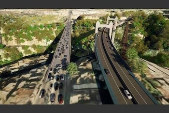 Vista aérea de dos puentes paralelos, uno con congestión vehicular y otro con pocos coches, flanqueados por vegetación y un puente ferroviario