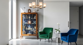 Rincón de vivienda con vitrina antigua de madera, dos sillones de terciopelo (verde y azul), lámpara Art Déco y orquídea. Paredes blancas, suelo pulido gris y luz natural de un ventanal.