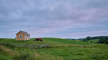 Casita de madera clara, tractor rojo, cerca de madera, colina verde, campo, cielo nublado.