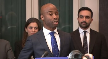 Un hombre negro calvo con traje oscuro y corbata azul habla en un podio. Un hombre con barba oscura está a su derecha y una mujer se ve parcialmente detrás