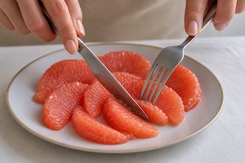 Dos manos sostienen un cuchillo y un tenedor para cortar gajos de pomelo rosado dispuestos en un plato redondo.