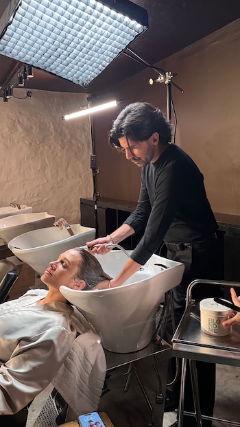 Max Jara, vestido de negro, lava el cabello de una mujer recostada en un lavacabezas blanco bajo grandes luces de estudio en un salón