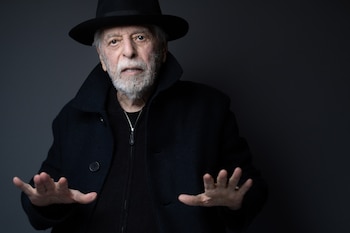 Hombre mayor con barba blanca, sombrero fedora negro y abrigo oscuro. Manos levantadas hacia adelante, palmas abiertas, sobre un fondo gris