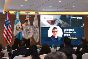 Mujer habla en un podio con logos de AmCham Guatemala, banderas de Estados Unidos y Guatemala, y una audiencia. Pantalla muestra retrato, texto de decreto y billetes