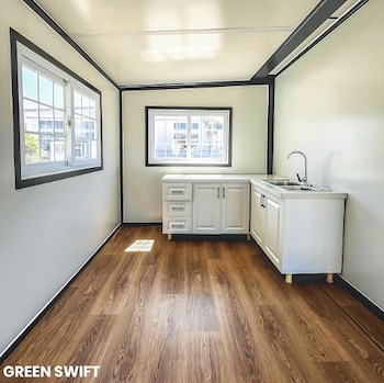 Interior de habitación con dos ventanas, piso de madera, paredes claras, gabinetes de cocina blancos con fregadero doble y grifo