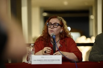 Mujer con cabello rojizo, gafas y camisa roja, habla por un micrófono azul. Una botella de agua y una placa de identificación están sobre la mesa, con fondo difuminado