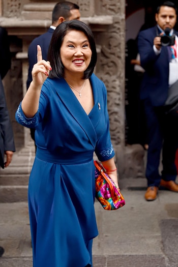 Mujer con vestido azul levanta el dedo índice y sonríe. Lleva collar de cruz, uñas rojas y cartera de colores. Hombres y muro de piedra en el fondo