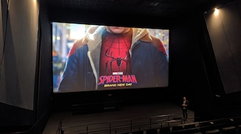 Una gran pantalla de cine proyecta el logo de Marvel Studios y el título Spider-Man: Brand New Day. Un hombre está de pie en el escenario, y se ven butacas vacías.