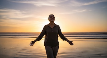Mujer sonriendo con ojos cerrados y brazos extendidos en una playa al atardecer. La luz cálida del sol poniente ilumina el mar y la orilla.