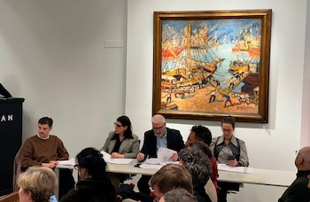 Varias personas sentadas en una mesa larga. Detrás, en una pared blanca, un cuadro enmarcado de un puerto con barcos. A la izquierda, un atril negro