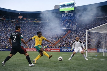 Un jugador de fútbol de Sudáfrica patea un balón hacia la portería, defendida por Oscar Pérez y Francisco Javier Aguilar, en un estadio lleno de aficionados.