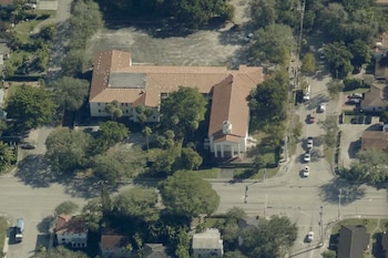 Vista aérea de una gran propiedad con edificios de techos rojizos y una fachada blanca en Miami, rodeada de árboles y calles con vehículos