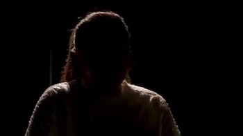 Primer plano de una persona, probablemente una mujer, en silueta oscura, con luz en el cabello y los hombros, sobre un fondo negro