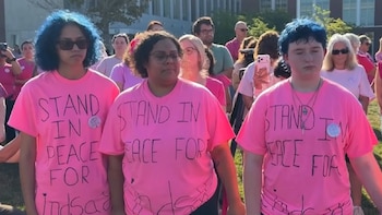 Tres personas al frente de una multitud llevan camisetas rosadas con letras escritas a mano en el torso