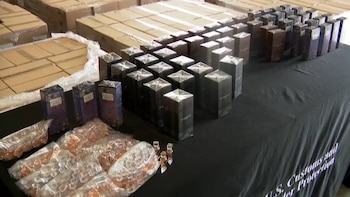 Una mesa negra muestra cajas de perfumes falsificados de marcas como Gucci y Burberry, junto a pequeñas botellas en bolsas transparentes. Al fondo, pilas de cajas de cartón