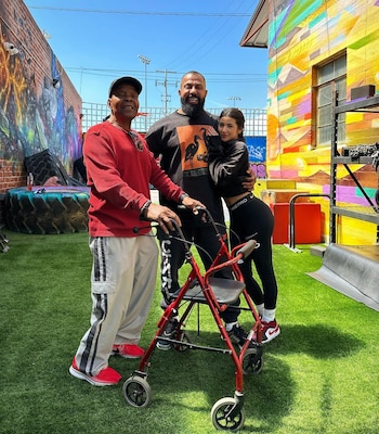 Un hombre mayor con camiseta roja y andador, flanqueado por un hombre barbudo y una mujer, sonríen en un patio con césped artificial y murales vibrantes