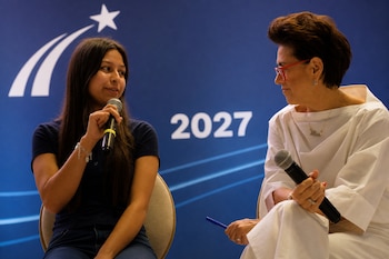 Dos mujeres sentadas con micrófonos conversan frente a una pared azul con el número 2027 y un logotipo blanco.