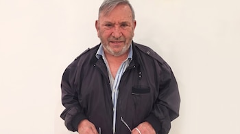 Hombre de edad avanzada con barba cana, chaqueta azul oscuro, camisa a rayas y anteojos frente a un fondo blanco.
