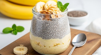 Vaso de natilla de chía y plátano con rodajas de plátano, coco tostado, canela y menta, sobre tabla de madera. Plátanos y semillas de chía al fondo.