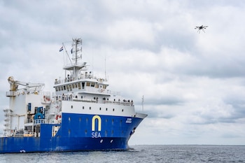 Buque de investigación blanco y azul N-Sea Geosea de la Armada Real de Países Bajos en el mar. Un dron vuela en el cielo nublado. Personas en cubierta, bandera neerlandesa