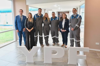 Seis personas, algunos en traje de negocios y otros en uniformes de trabajo, posan sonrientes detrás de un gran letrero blanco de YPF en un interior luminoso