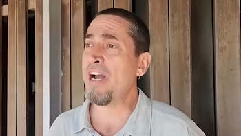Primer plano de un hombre con barba de chivo, camisa clara, hablando con la boca abierta, frente a un fondo de listones de madera verticales