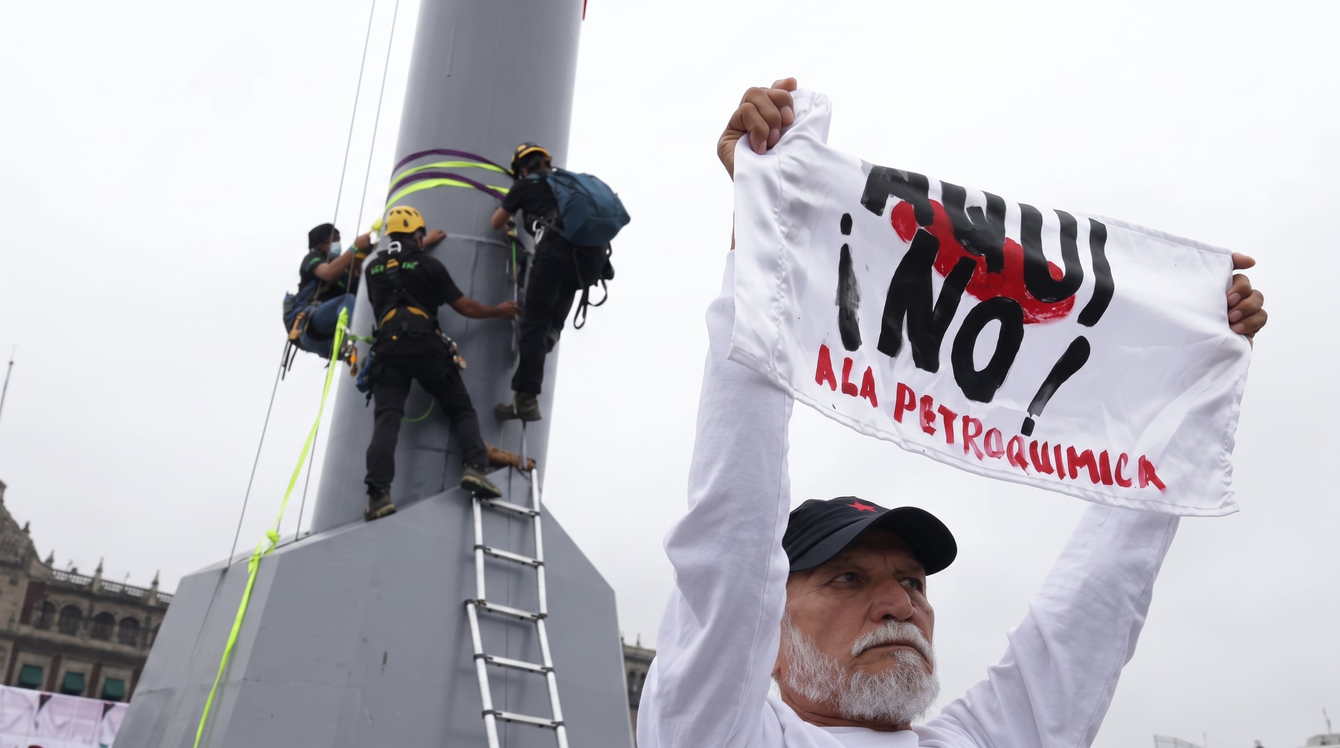 Un manifestante sostiene una pancarta en rechazo a la petroquímica mientras varias personas con equipo de seguridad escalan una asta bandera en una plaza pública. (Imagen Ilustrativa Infobae)