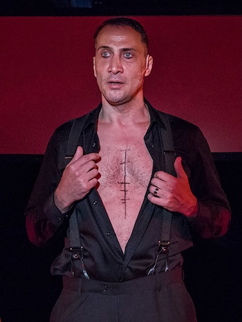 Primer plano de Luciano Cáceres con una camisa negra abierta, tirantes y una línea vertical con marcas dibujada en su pecho, mirando hacia arriba