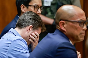 Un hombre caucásico con cabello castaño y barba corta, vestido con una camisa azul, se cubre el rostro con una mano, sentado en un tribunal junto a otras personas