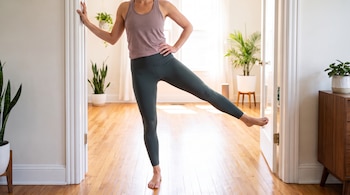 Una persona con camiseta y leggings realiza un ejercicio de pierna, apoyada en el marco de una puerta, en una sala con piso de madera y plantas.