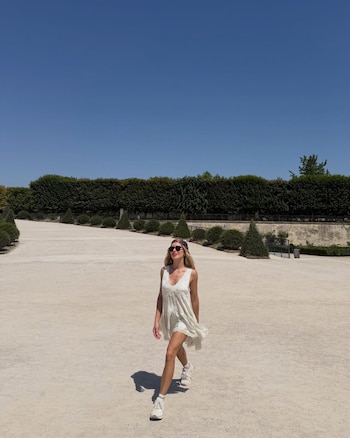 Una mujer rubia con lentes de sol, pañuelo en la cabeza, vestido blanco y zapatillas camina en un sendero de tierra en un parque con setos altos