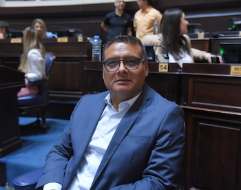 Luis Ontiveros, un hombre con gafas y traje azul, sentado en un asiento azul en una sala con mesas de madera y otras personas al fondo