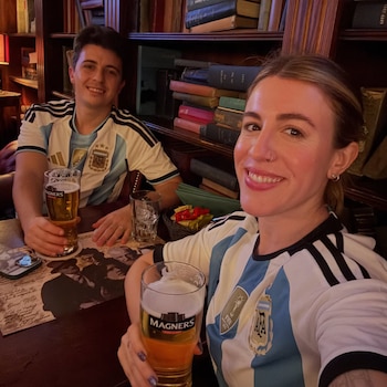 Un hombre y una mujer, ambos con camisetas de la selección argentina, brindan con vasos de cerveza en una mesa de un bar