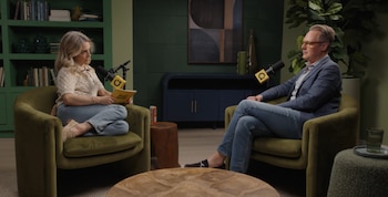 Hombre de mediana edad con gafas, chaqueta azul y camisa a cuadros, sentado en un sillón verde. Un micrófono negro con logo amarillo. Plantas y cuadro abstracto en fondo
