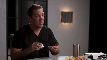 Un hombre caucásico de cabello corto y claro, el Dr. Alex Tatem, con uniforme quirúrgico negro, habla sentado en una mesa, sosteniendo un pequeño frasco y gesticulando