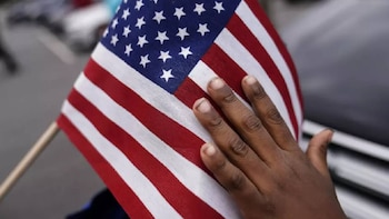 Primer plano de una mano de piel oscura tocando una pequeña bandera de Estados Unidos. La bandera tiene estrellas blancas sobre azul y franjas rojas y blancas, y está en un mástil de madera